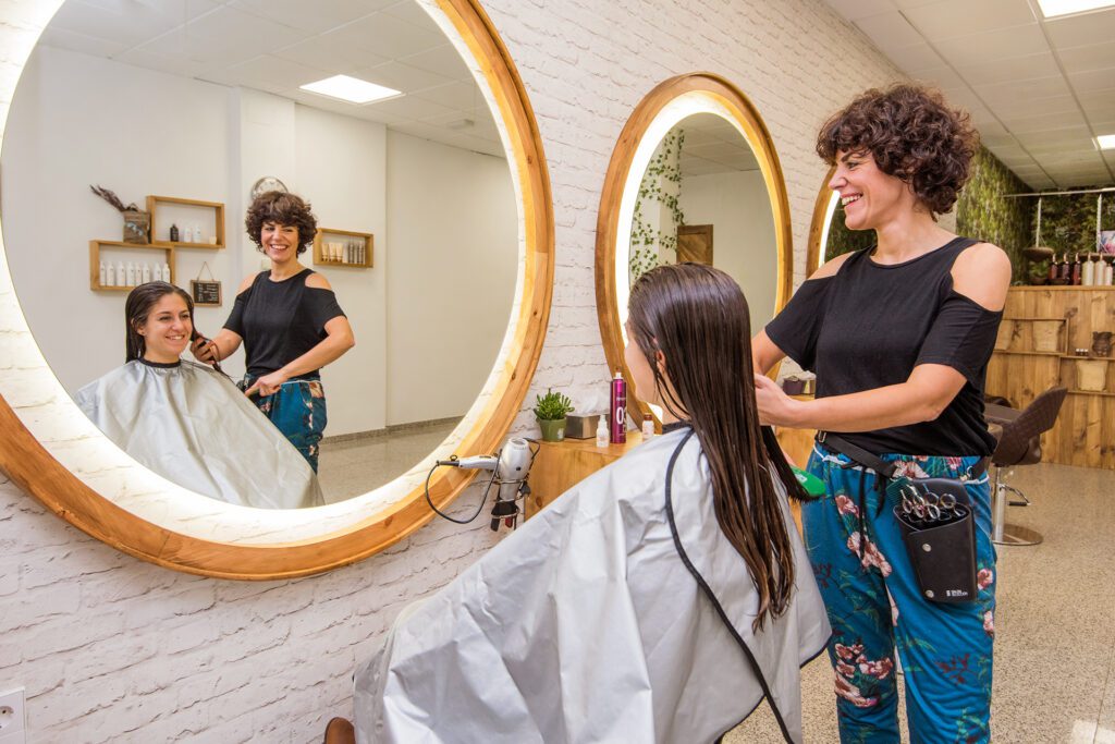 Een glimlachende kapster met kort krullend haar borstelt het lange, vochtige haar van een klant voor een grote, ronde, verlichte spiegel. De scène wordt weerspiegeld in de spiegel, die het interieur van de salon toont.