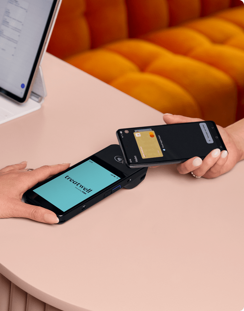 Een persoon houdt een zwarte betaalterminal vast met het lichtblauwe scherm van het merk "Treatwell", terwijl een andere persoon diens smartphone ertegenaan houdt om een contactloze betaling te doen via een digitale portemonnee. De scène speelt zich af op een lichtroze tafel.