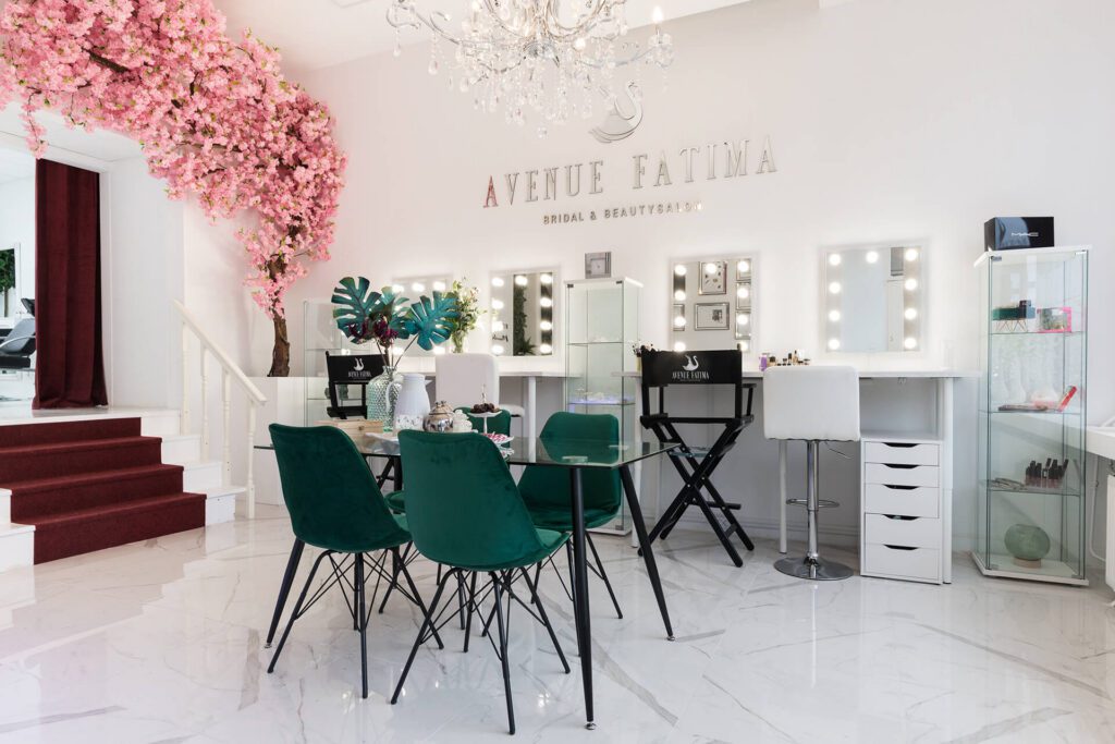 Een lichte, elegante bruids- en schoonheidssalon genaamd "Avenue Fatima". De ruimte beschikt over meerdere make-upstations met verlichte spiegels, smaragdgroene fluwelen stoelen rond een glazen tafel en een decoratieve roze kersenbloesemboom.