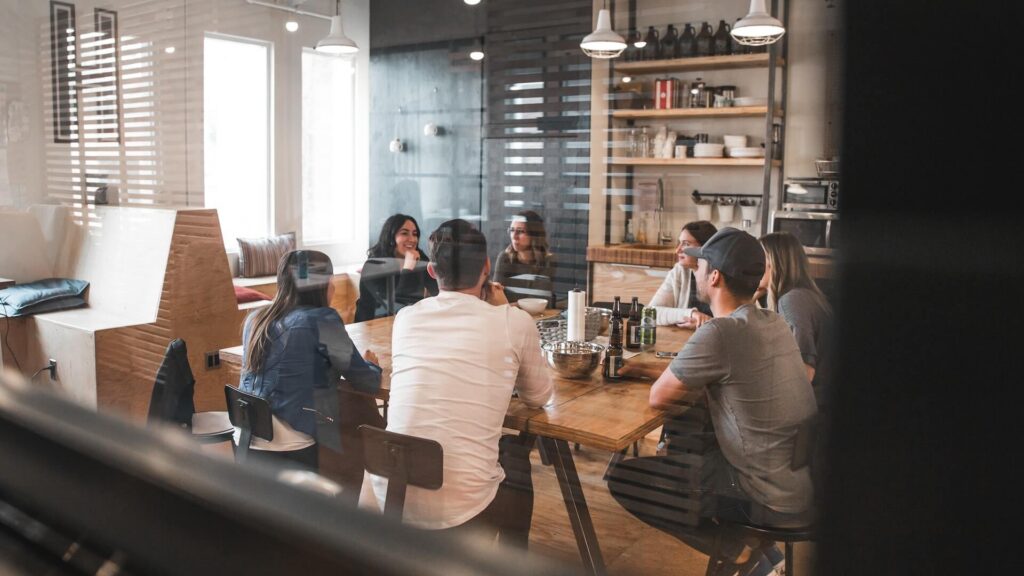 Een spontane blik door een glazen ruit naar een groep collega's die rond een lange houten tafel zitten in een moderne open kantoorkeuken of kantine, pratend en met drankjes.